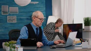 Senior man using laptop and tablet in same time working in office living room at project. Busy focused old employee writing, searching, analysing reports while elderly wife watching tv in background video