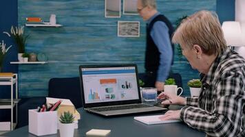 Focused senior entrepreneur checking graphics from laptop writing on notebook working from business office overtime. Busy employee using modern technology network wireless reading typing, searching video
