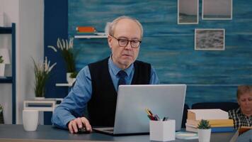 Senior businessman reading reports sitting in front of laptop working from home drinking coffee. Retired man using modern technology analysing typing searching while wife sitting on couch reading a book video