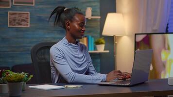 Black woman typing on computer and writing list of tasks on sticky notes putting it on laptop sitting on desk in living room workplace late at night. African freelancer working from home overtime video