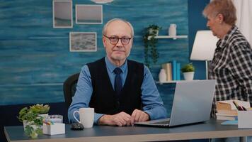 Elderly successful businessman entrepreneur sitting in workspace looking at camera smiling. Senior manager working from home for start up business, prepared for online meeting using moden gadgets video
