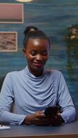 Vertical . African lady taking break typing on smartphone, reading news smiling sitting at desk in living room late at night. Employee using modern technology network wireless doing overtime for job searching video