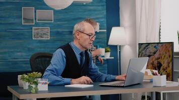 multitasking pensionato uomo lettura su il computer portatile e scrittura su taccuino Lavorando a partire dal casa. focalizzata vecchio a distanza dipendente ricerca, analizzando finanziario rapporti mentre anziano moglie Guardando tv nel sfondo video