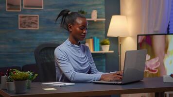 Black woman typing on computer smiling and writing informations of company on notebook sitting on desk in living room workplace late at night. African freelancer working from home overtime at laptop. video