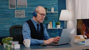 Focused old entrepreneur with headphones listening music while checking graphics writing on laptop working from home. Retired man using modern technology reading typing, searching on computer video