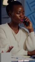 African employee speaking at phone while working at laptop late at night. Busy focused freelancer doing overtime for job reading writing, searching, respecting deadline of financial project, overworking video