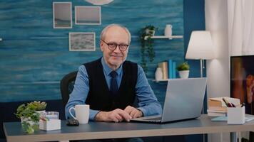 Senior man looking at camera smiling sitting in workplace at desk working from home while old wife watching tv in background. Retired business person prepared for online meeting using moden gadgets video