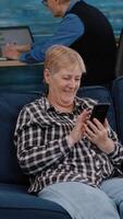 Vertical . Middle aged woman relaxing holding smartphone reading e book sitting on sofa at home, while senior adult man working at laptop in background. Person enjoying using notepad browsing internet shopping video
