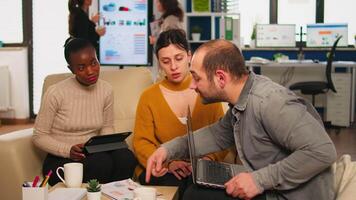 Confident company manager giving working tasks to diverse teammates sitting on couch in start up office. Multiethnic team discussing project ideas at brainstorming meeting using digital devices video