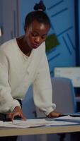 Stressed african manager woman working with financial documents standing at desk checking graphs, holding papers, reading raports late at night in start-up office doing overtime to respect deadline. video
