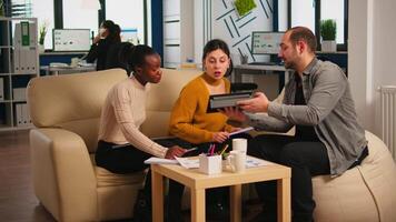 Multiethnic team analysing informations from tablet discussing sitting on couch brainstorming on new project for start up company. Diverse businesspeople analysing financial reports during meeting. video