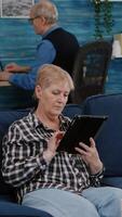 Vertical . Happy old senior grandmother holding digital tablet browsing, searching informations, shopping online, while adult man working at laptop in background. Person enjoying using notepad gadget video