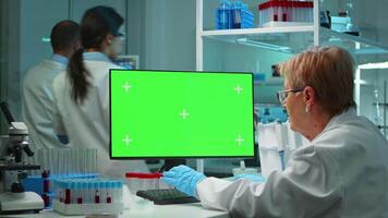 Senior doctor working at computer with green screen in modern equipped lab late at night. Team of microbiologists doing vaccine research writing on device with chroma key, isolated, mockup display. video