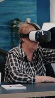 Vertical . Retired woman experiencing virtual reality using vr headset in living room working from home. Old remote employee searching, analysing financial reports while elderly wife watching tv in background video