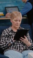 Vertical . Middle aged old woman relaxing holding tablet reading e book sitting on sofa at home, while senior adult man working at laptop in background. Person enjoying using notepad browsing internet shopping video