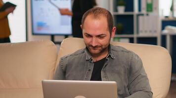 Manager sitting on couch in front of camera smiling while multiethnic team working in background with financial reports. Diverse coworkers talking about startup financial company in modern office. video