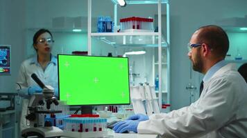 Doctor working at computer with green screen over time sitting in modern equipped lab. Team of microbiologists doing vaccine research writing on device with chroma key, isolated, mockup display. video