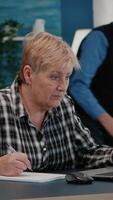 Vertical . Old woman entrepreneur checking financial project of company looking on laptop analysing annual statistics data, taking notes on notebook sitting at desk in living room office working from home. video