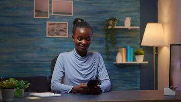 African lady taking break typing on smartphone, reading news smiling sitting at desk in living room late at night. Employee using modern technology network wireless doing overtime for job searching video