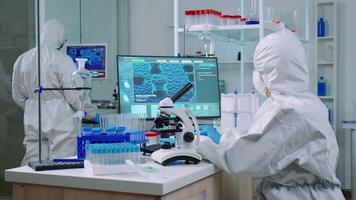 Chemist in protection suit typing on pc and analyzing virus sample using microscope in scientific lab. Team of doctors examining vaccine evolution using high tech researching diagnosis against covid19 video