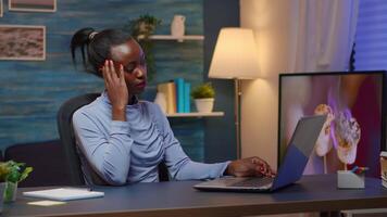 Tired black businesswoman working overtime reading projects sitting at desk in living room using laptop. Busy focused employee using modern technology network wireless overworking for job searching video