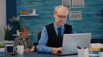 Retired manager sitting at desk in front of camera smiling after typing at laptop working from home while senior wife reading a book in background. Man prepared for online meeting using moden gadgets video