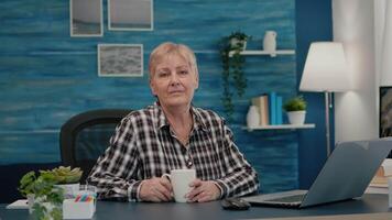 Senior focused woman looking at camera sitting in workplace at desk working from home while old husband watching tv in background. Retired business person prepared for online meeting using moden gadgets video