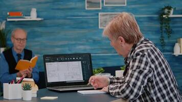 Senior woman architect analysing digital prototype with plans from laptop working from home, taking notes on notebook. Elderly designer using cad software to create a 3D concept of buildings video