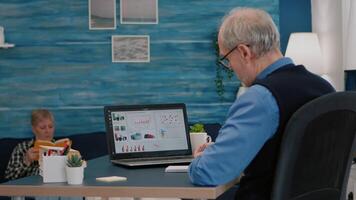 Middle aged senior older man e-learning online concept, watching business graphs, charts on laptop computer remote working, social distance learning, writing noteson notebook from home office. video