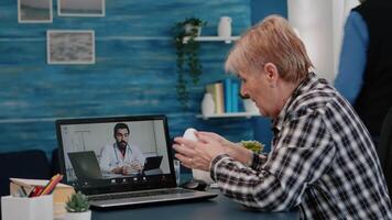 Old woman sitting at table at home looking at laptop answering question about health talks with therapist by videoconference call application, holding pills flask Remote medical consultation concept video
