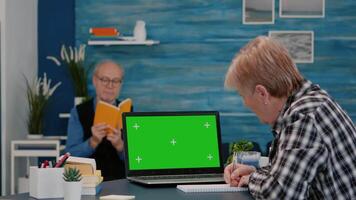 Senior woman looking at green screen monitor working from home, taking notes on notebook. Elderly businesswoman watching desktop laptop display with green mockup, chroma key, while man reading a book video