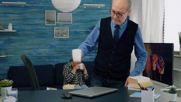 Retired project manager working from home, opening laptop reading news, typing and analysing financial statistics, while his wife drinking tea in background. Old man using modern technology for job video