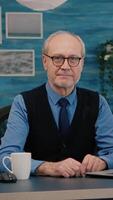 Vertical . Elderly successful businessman entrepreneur sitting in workspace looking at camera smiling. Senior manager working from home for start up business, prepared for online meeting using moden gadgets video