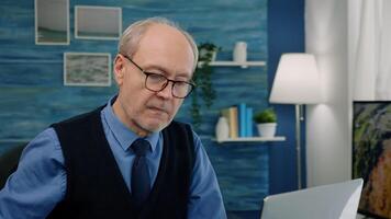 Close up of senior man writing at laptop working from home analysing financial project. Elegant retired employee with eyeglasses using modern technology reading typing on computer video