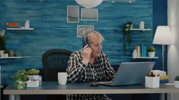 Happy old woman entrepreneur with headset listening music while checking statistics, writing on laptop working from home. Retired worker using modern technology reading typing, searching on computer video