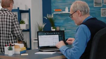 Senior man architect analysing digital prototype with plans from laptop working from home, taking notes on notebook. Elderly engineer designer using cad software to create a 3D concept of buildings. video
