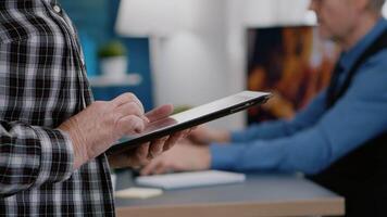 Close up of elderly woman employee texting, reading messages from tablet standing in home workspace while man working at laptop in background. Closeup of person hands holding notepad analysing graphs video