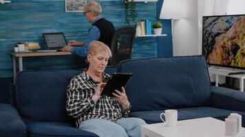 Happy old senior grandmother holding digital tablet browsing, searching informations, shopping online, while adult man working at laptop in background. Person enjoying using notepad gadget video