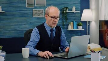 Tired elderly man sitting at desk and taking a sip of coffee, looking at laptop working from home workspace while his wife is reading a book in background. Focused old entrepreneur checking graphics video