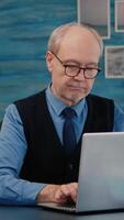 Vertical . Retired manager sitting at desk in front of camera smiling after typing at laptop working from home while senior wife reading a book in background. Man prepared for online meeting using moden gadgets video