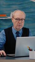Vertical . Senior businessman reading reports sitting in front of laptop working from home drinking coffee. Retired man using modern technology analysing typing searching while wife sitting on couch reading a book video