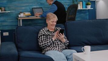 Middle aged woman relaxing holding smartphone reading e book sitting on sofa at home, while senior adult man working at laptop in background. Person enjoying using notepad browsing internet shopping video