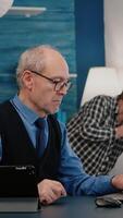 Vertical . Senior man using laptop and tablet in same time working in office living room at project. Busy focused old employee writing, searching, analysing reports while elderly wife watching tv in background video