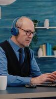 Vertical . Focused old entrepreneur with headphones listening music while checking graphics writing on laptop working from home. Retired man using modern technology reading typing, searching on computer video
