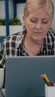 Vertical . Old focused woman looking at camera after typing on laptop sitting in workplace at desk working from home while husband watching tv in background. Retired business person prepared for online meeting video