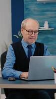 Vertical . Happy old manager receving good news on laptop working from home for startup company, gesturing victory. Retired employee using modern technology reading typing searching while senior wife watching TV video