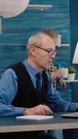Vertical . Multitasking retired man reading on laptop and writing on notebook working from home. Focused old remote employee searching, analysing financial reports while elderly wife watching tv in background video