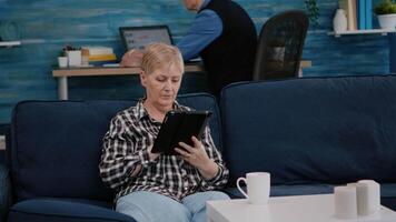 Middle aged old woman relaxing holding tablet reading e book sitting on sofa at home, while senior adult man working at laptop in background. Person enjoying using notepad browsing internet shopping video