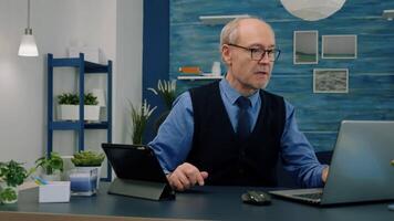 Elderly man using tablet and laptop in same time compering financial graphs working from home sitting in workplace. Busy focused old employee writing, searching, analysing reports video
