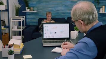 Tired elderly entrepreneur checking graphics from laptop writing on notebook working from business office overtime. Busy employee using modern technology network wireless reading typing, searching video